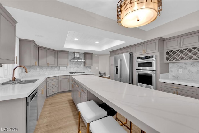 kitchen featuring gray cabinetry, backsplash, wall chimney exhaust hood, a tray ceiling, and stainless steel appliances