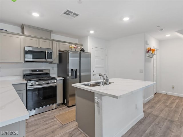 kitchen with gray cabinetry, light stone countertops, sink, stainless steel appliances, and a center island with sink