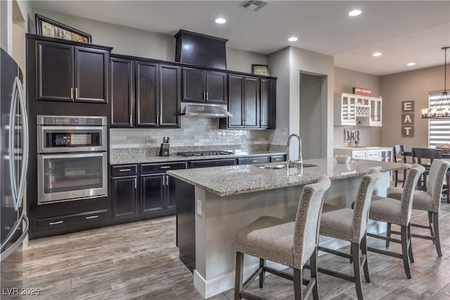 kitchen with light stone countertops, appliances with stainless steel finishes, a kitchen island with sink, sink, and decorative light fixtures