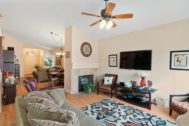 living room with a tiled fireplace, ceiling fan with notable chandelier, vaulted ceiling, and light wood-type flooring