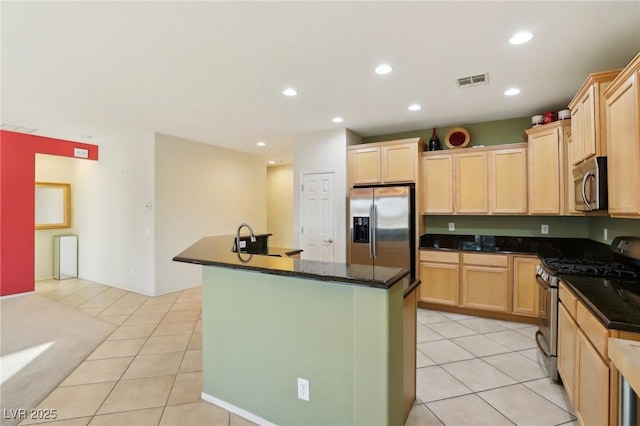 kitchen featuring dark stone counters, stainless steel appliances, a kitchen island with sink, light tile patterned floors, and light brown cabinets