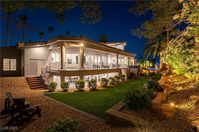 back of house at night featuring stucco siding, stairway, a chimney, and a lawn