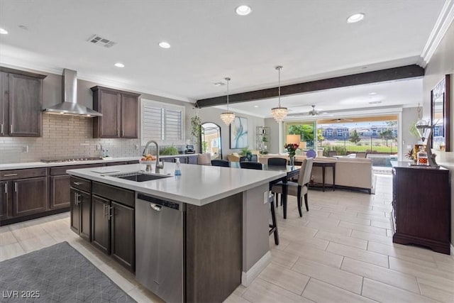 kitchen with sink, wall chimney exhaust hood, stainless steel appliances, beamed ceiling, and a center island with sink