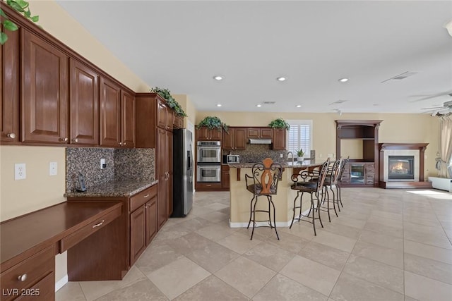 kitchen with tasteful backsplash, stainless steel appliances, a center island with sink, dark stone countertops, and a breakfast bar area