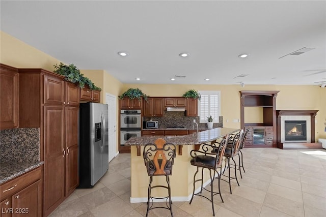 kitchen featuring dark stone counters, a center island with sink, decorative backsplash, appliances with stainless steel finishes, and a kitchen bar