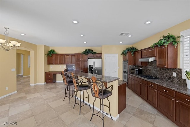 kitchen with decorative light fixtures, stainless steel appliances, dark stone counters, and an island with sink