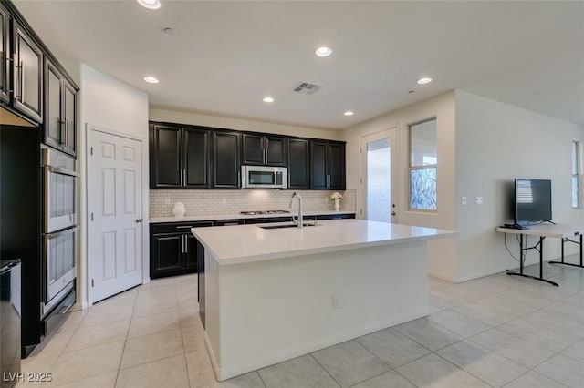 kitchen featuring backsplash, stainless steel appliances, sink, light tile patterned floors, and a center island with sink