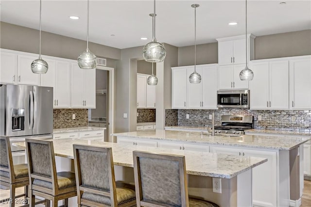 kitchen featuring light stone countertops, appliances with stainless steel finishes, decorative light fixtures, white cabinets, and an island with sink