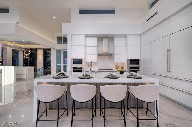 kitchen with double oven, a large island with sink, white cabinets, and wall chimney range hood