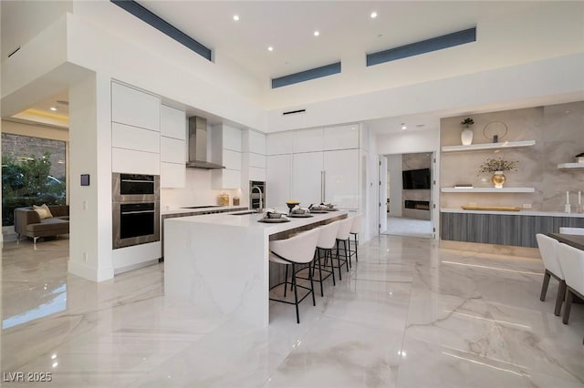 kitchen with a kitchen bar, stainless steel double oven, a kitchen island with sink, wall chimney range hood, and white cabinets