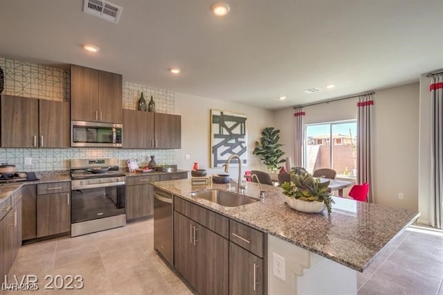 kitchen featuring a kitchen island with sink, sink, appliances with stainless steel finishes, tasteful backsplash, and light stone counters