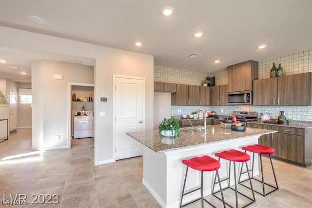 kitchen featuring a breakfast bar, stainless steel appliances, light stone counters, washer / clothes dryer, and a center island with sink