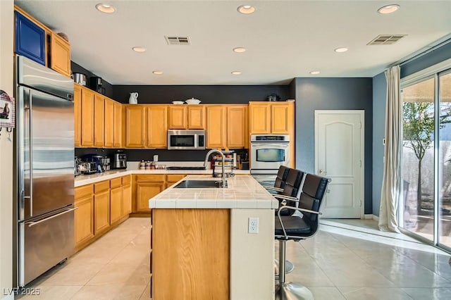 kitchen featuring sink, a center island with sink, light tile patterned floors, appliances with stainless steel finishes, and tile counters