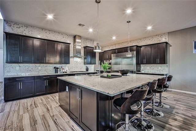 kitchen with a center island with sink, wall chimney range hood, appliances with stainless steel finishes, decorative light fixtures, and dark brown cabinets