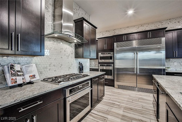 kitchen featuring light stone counters, dark brown cabinets, wall chimney range hood, and appliances with stainless steel finishes