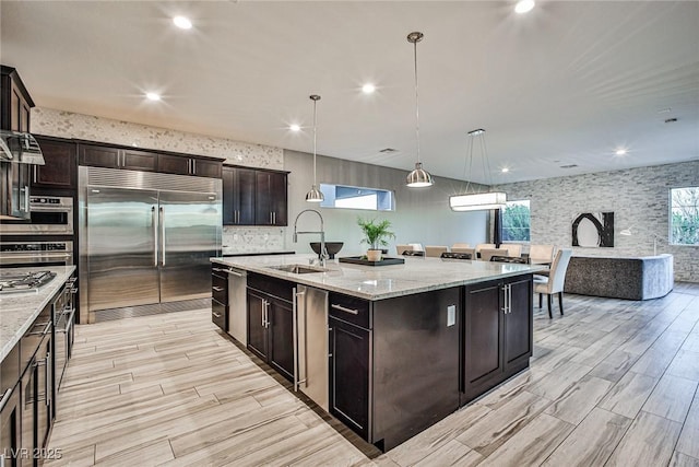 kitchen with sink, built in fridge, light stone counters, pendant lighting, and a center island with sink