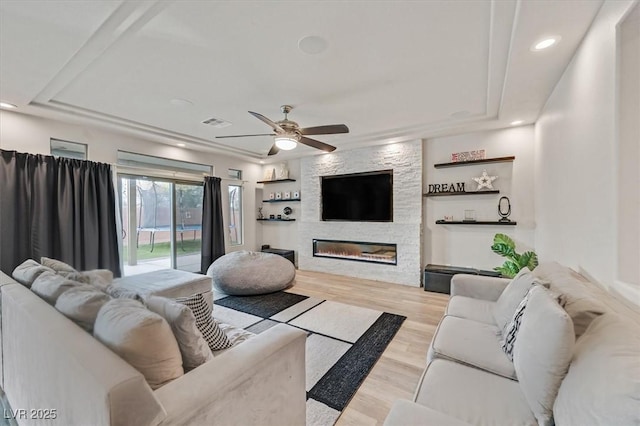 living room with light wood-type flooring, a tray ceiling, a stone fireplace, and ceiling fan