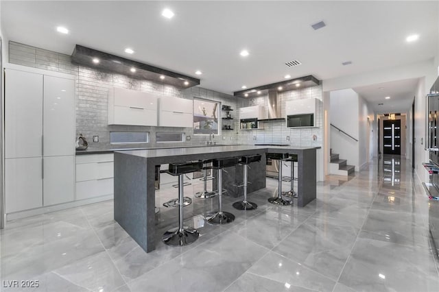 kitchen featuring a kitchen bar, backsplash, wall chimney exhaust hood, a spacious island, and white cabinetry