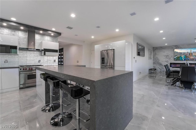 kitchen with a breakfast bar area, white cabinetry, high end appliances, a kitchen island, and wall chimney exhaust hood