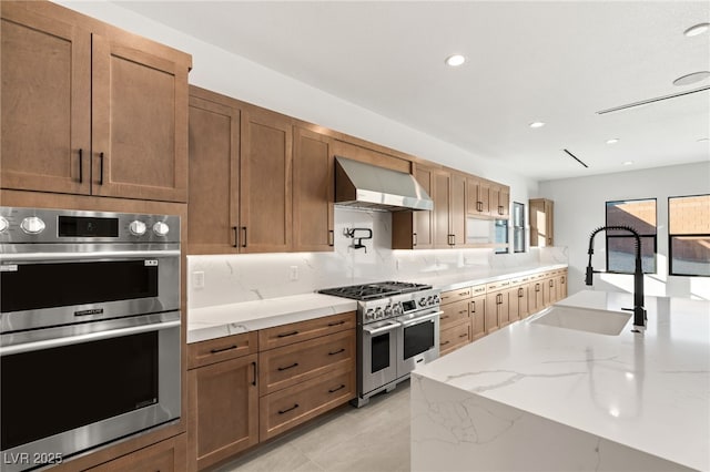 kitchen featuring sink, stainless steel appliances, wall chimney range hood, light stone counters, and backsplash