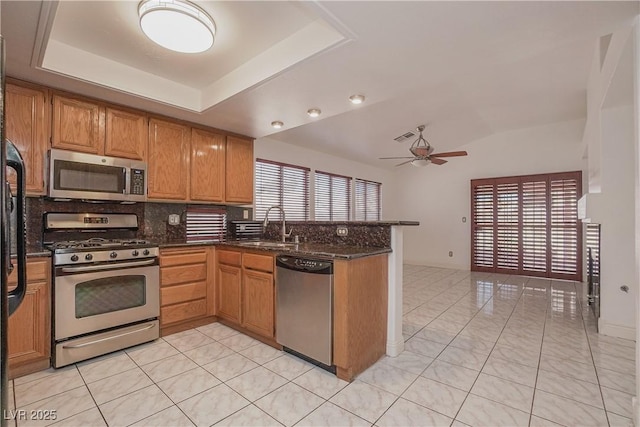 kitchen with kitchen peninsula, stainless steel appliances, a raised ceiling, ceiling fan, and dark stone countertops