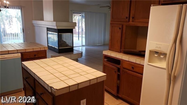 kitchen with a tile fireplace, dishwasher, tile counters, white fridge with ice dispenser, and a kitchen island