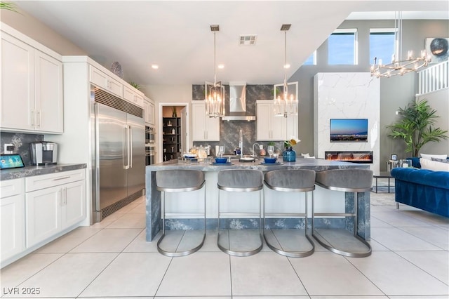 kitchen with wall chimney exhaust hood, decorative light fixtures, tasteful backsplash, built in fridge, and white cabinetry