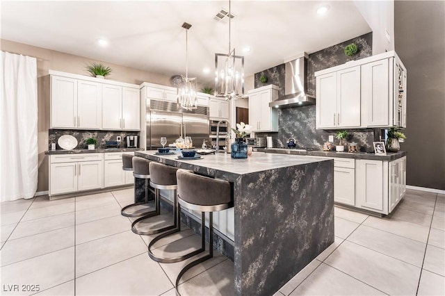 kitchen with wall chimney exhaust hood, decorative light fixtures, a kitchen island with sink, white cabinetry, and appliances with stainless steel finishes