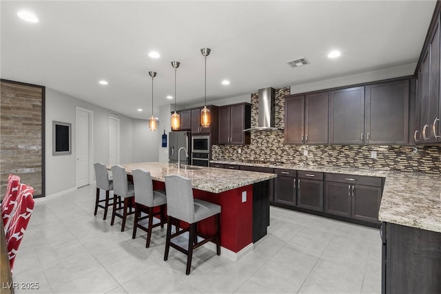 kitchen with a center island with sink, a kitchen breakfast bar, wall chimney exhaust hood, decorative light fixtures, and light stone counters