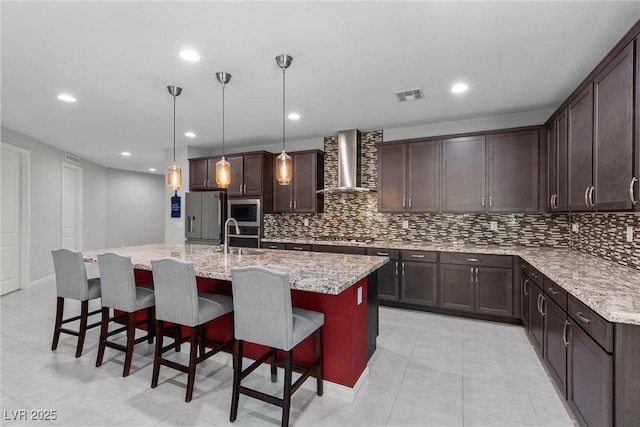 kitchen featuring a kitchen island with sink, wall chimney range hood, light stone countertops, decorative light fixtures, and stainless steel appliances