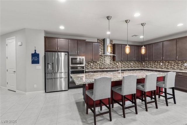 kitchen featuring wall chimney exhaust hood, dark brown cabinets, stainless steel appliances, decorative light fixtures, and an island with sink