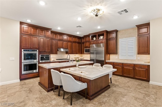 kitchen with a kitchen bar, built in appliances, light stone countertops, and a center island with sink