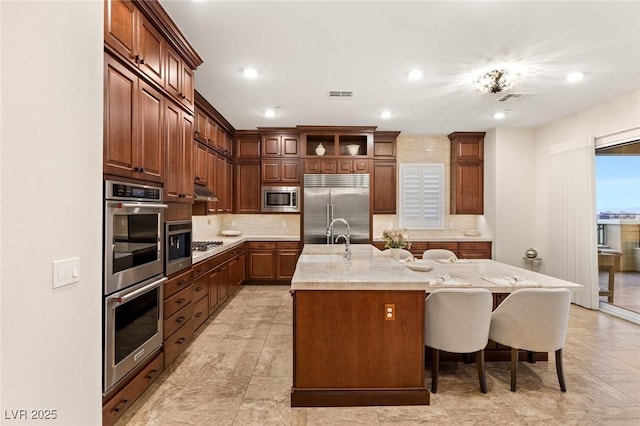 kitchen with a kitchen breakfast bar, light stone counters, built in appliances, an island with sink, and decorative backsplash