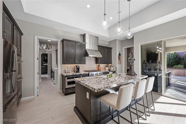 kitchen with backsplash, wall chimney exhaust hood, an island with sink, appliances with stainless steel finishes, and light stone counters