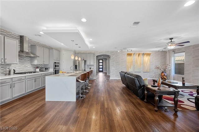 kitchen featuring appliances with stainless steel finishes, wall chimney exhaust hood, a kitchen island with sink, pendant lighting, and gray cabinets