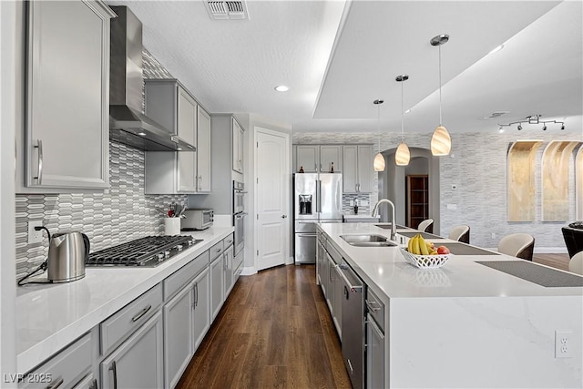 kitchen featuring gray cabinetry, stainless steel appliances, wall chimney range hood, a center island with sink, and hanging light fixtures