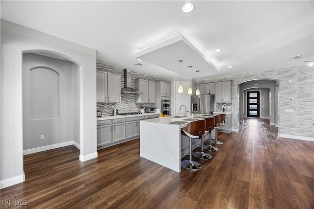 kitchen featuring gray cabinetry, pendant lighting, a kitchen island with sink, wall chimney exhaust hood, and stainless steel appliances