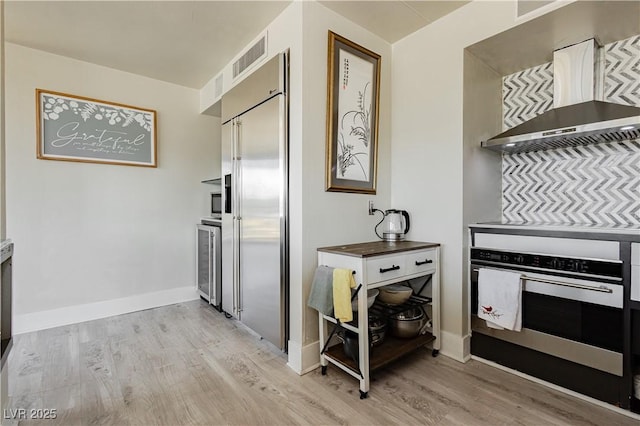 kitchen featuring light wood-type flooring, wall chimney exhaust hood, oven, stainless steel built in fridge, and wine cooler