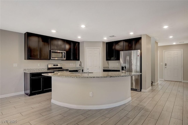 kitchen featuring light stone counters, dark brown cabinetry, stainless steel appliances, a kitchen island with sink, and sink