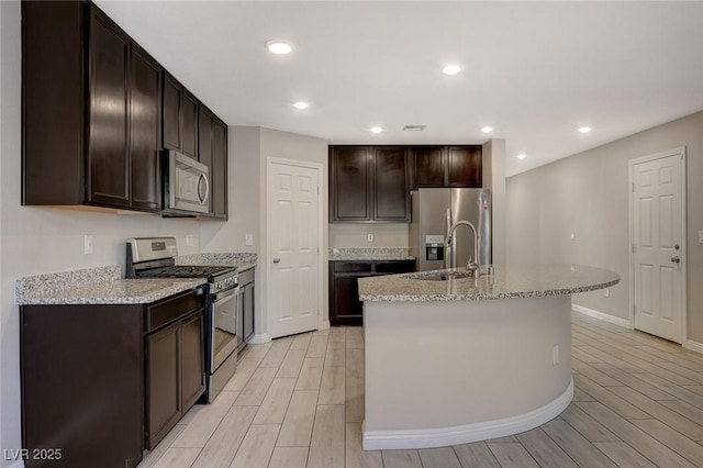 kitchen featuring light stone countertops, dark brown cabinets, stainless steel appliances, a kitchen island with sink, and sink