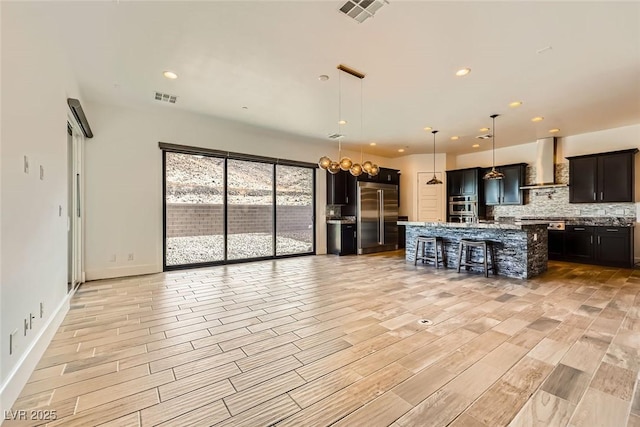 kitchen featuring pendant lighting, stainless steel built in refrigerator, a kitchen island with sink, light stone countertops, and wall chimney range hood