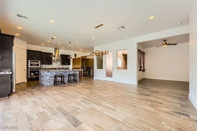 kitchen featuring wall chimney exhaust hood, a breakfast bar area, decorative light fixtures, a center island with sink, and decorative backsplash