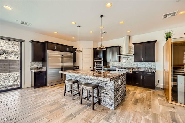 kitchen with decorative light fixtures, a kitchen island with sink, light stone counters, stainless steel appliances, and wall chimney range hood