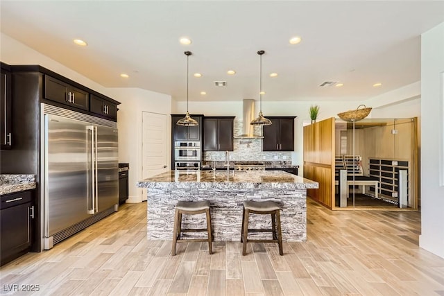 kitchen with stainless steel built in refrigerator, light stone countertops, a kitchen island with sink, and wall chimney exhaust hood