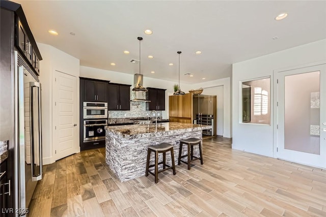 kitchen with wall chimney range hood, appliances with stainless steel finishes, hanging light fixtures, light stone countertops, and an island with sink