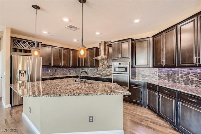 kitchen with an island with sink, pendant lighting, wall chimney range hood, and appliances with stainless steel finishes