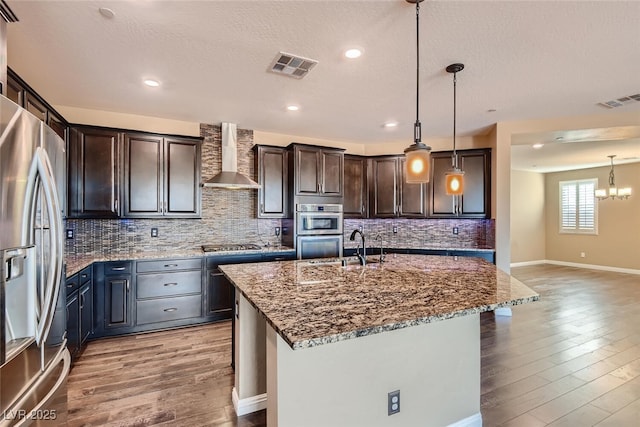 kitchen with appliances with stainless steel finishes, wall chimney exhaust hood, a kitchen island with sink, stone counters, and hanging light fixtures