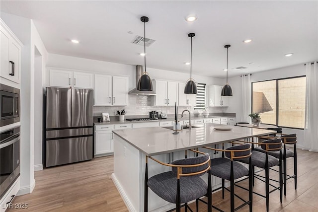 kitchen with appliances with stainless steel finishes, pendant lighting, wall chimney exhaust hood, and an island with sink