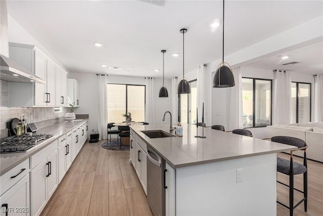 kitchen featuring appliances with stainless steel finishes, wall chimney range hood, decorative light fixtures, white cabinetry, and an island with sink