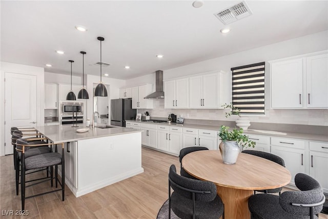 kitchen featuring hanging light fixtures, wall chimney range hood, a center island with sink, white cabinets, and appliances with stainless steel finishes